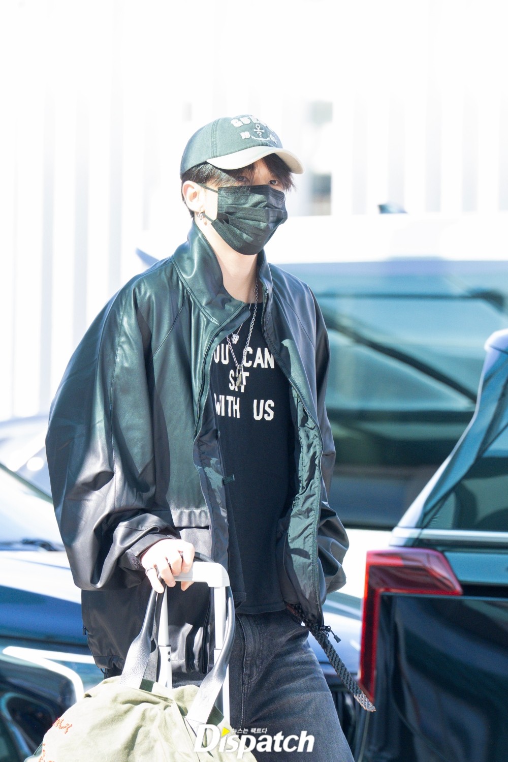 Hyunjin walking beside a car in an open black jacket with a printed shirt and suitcase.